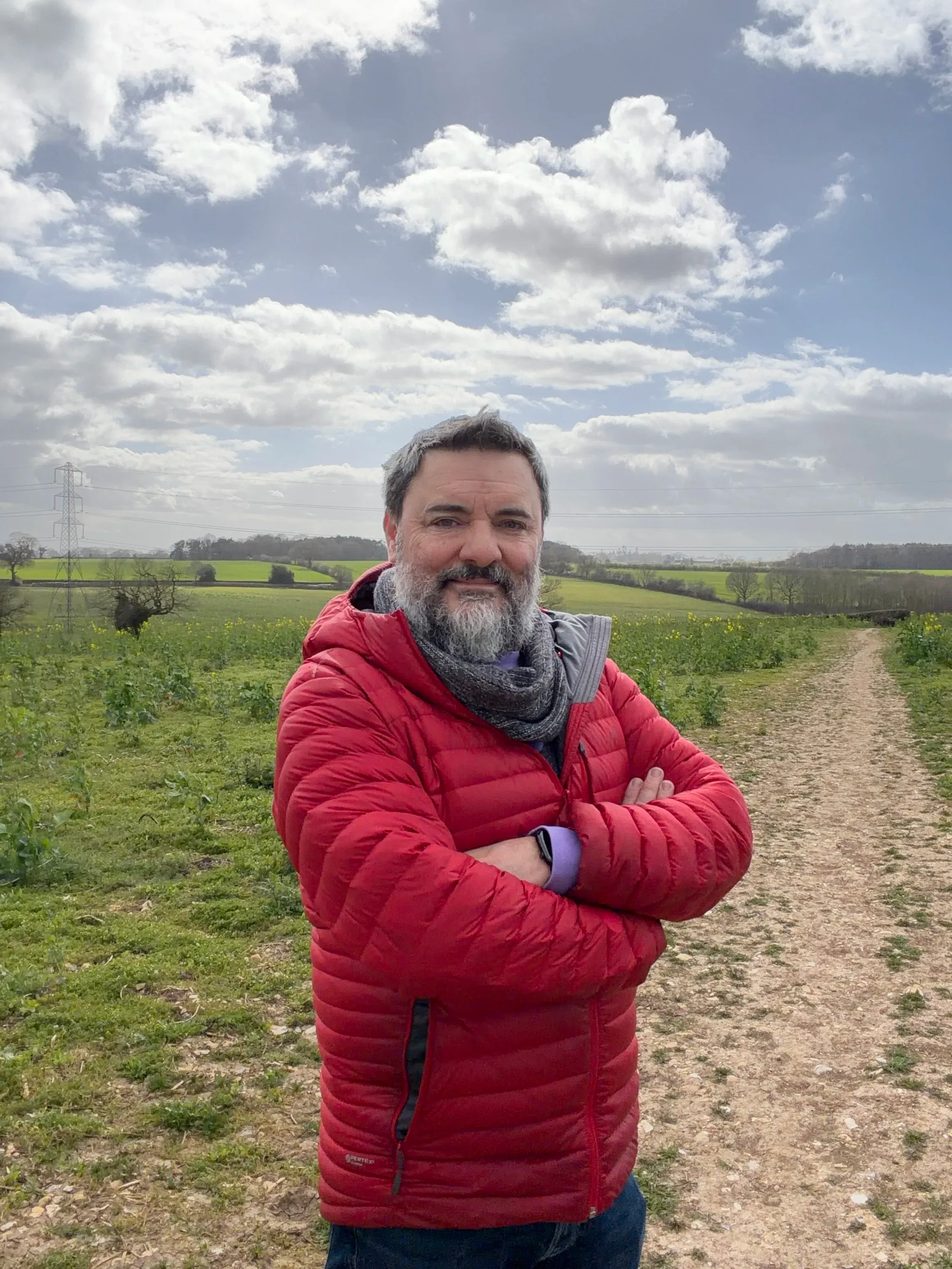 Chris Goor on a footpath in a red quilted jacket, arms folded, big clouded sky behind him