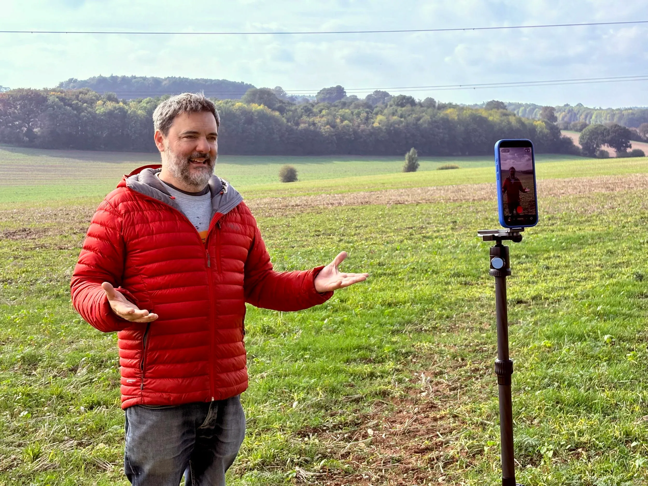 Chris Goor in a red jacket in an open field, arms out, talking to a phone on a tripod