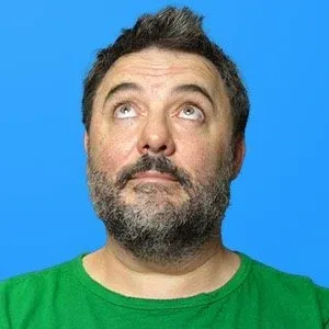 Chris Goor in a green T-shirt against a blue wall, looking up with a wry expression