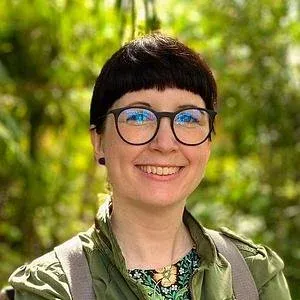 Christy, dark bob and glasses, smiling in front of green foliage